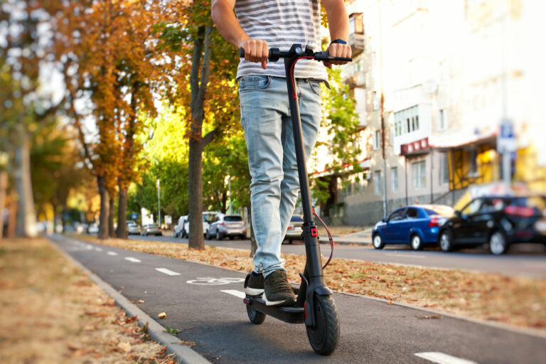 Person rides an electric scooter along a tree-lined city street in autumn leaves.