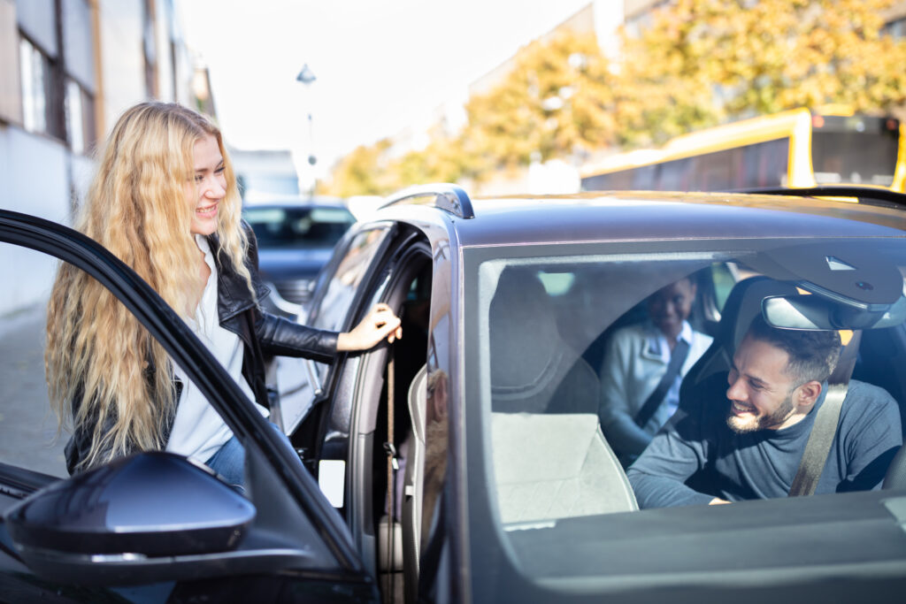 Woman with long blonde hair smiles as she opens the driver's door of a dark car on a city street, a man sits inside and smiles at her.
